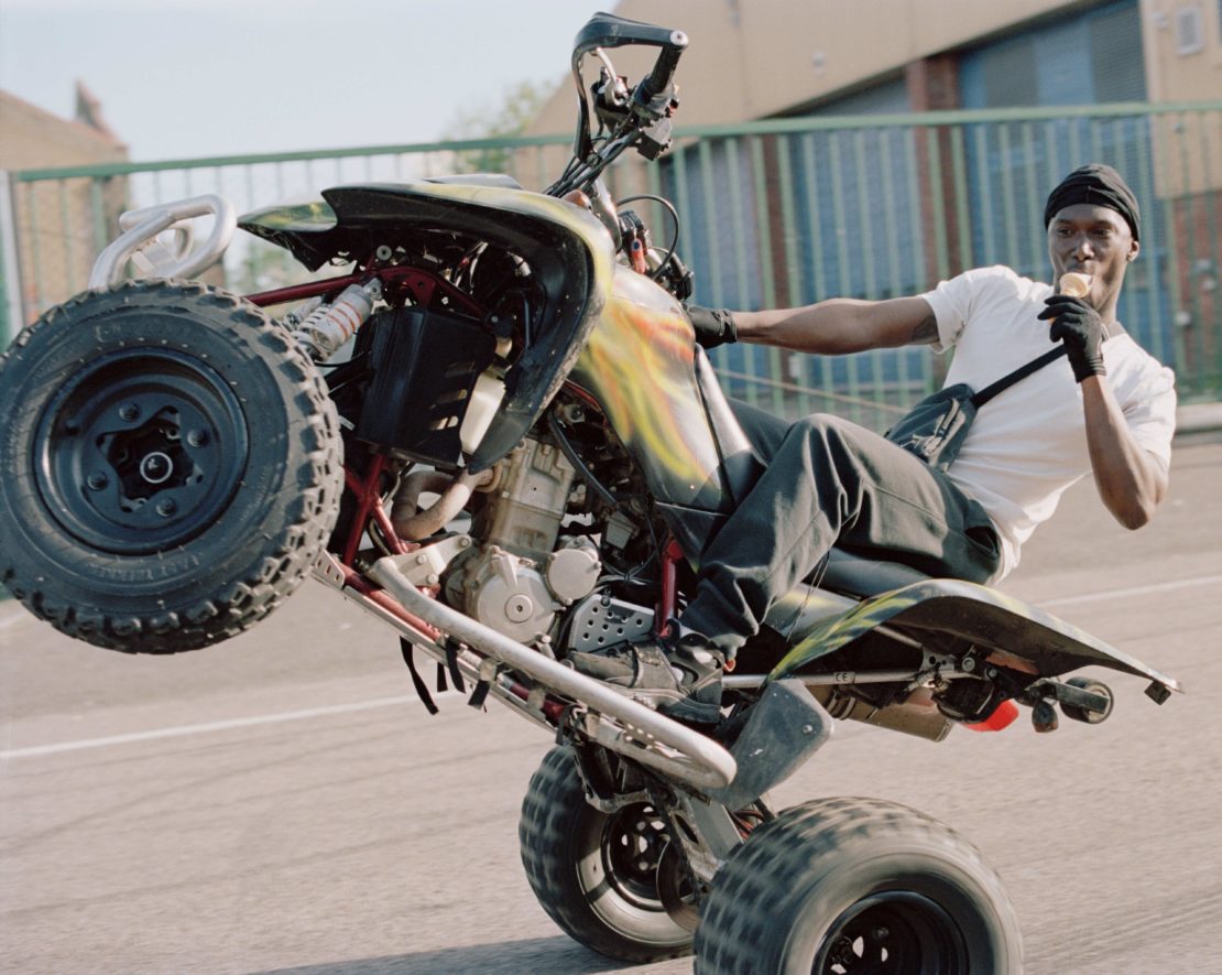 A black man doing a wheelie on a yellow and black ATV while eating, outdoors on pavement with green buildings in the background.