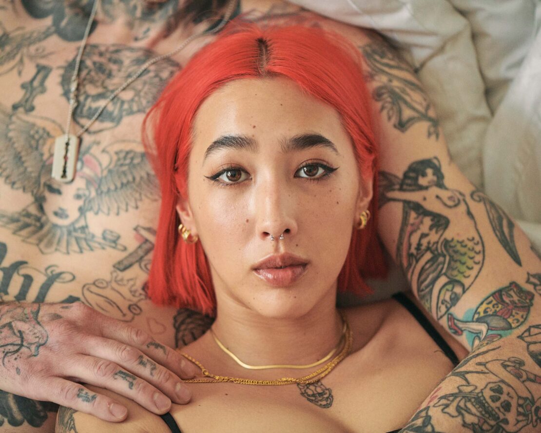 Close-up portrait of a woman with bright red hair lying on a bed, with a tattooed person in the background.