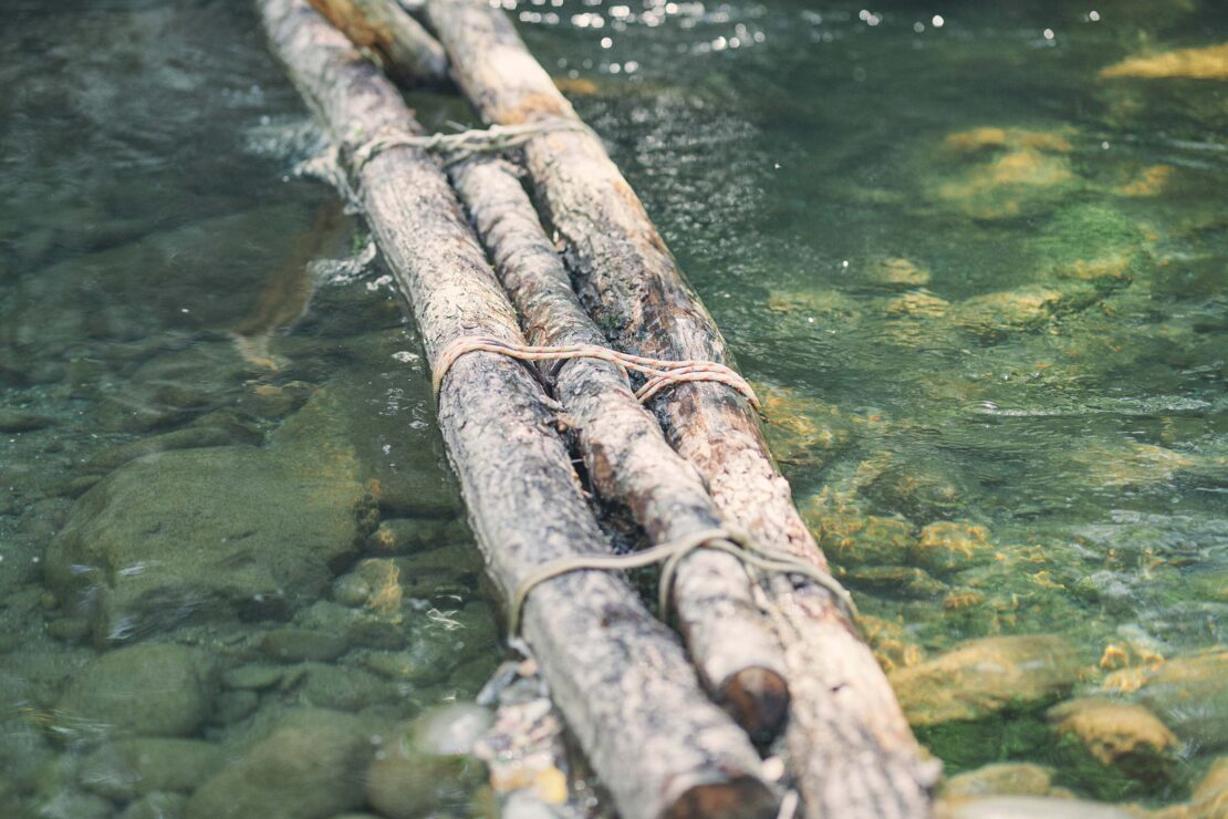Logs tied together form a makeshift bridge over a rocky riverbed, sparkling with sunlight and lush vegetation on its banks.