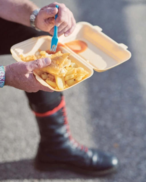 Adult enjoys fish and chips from a takeout container, using a blue fork on a sunny day, with black boots visible in the background.
