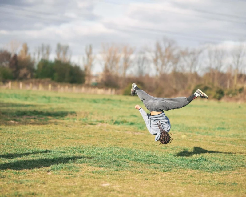 Teenage athlete performs a backflip on a grassy field under a cloudy sky, showcasing skill and agility in an outdoor environment.