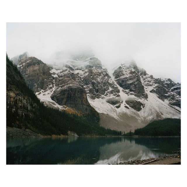 Scenic landscape of snow-capped mountains reflecting in a still lake under a cloudy sky, surrounded by a lush green forest.