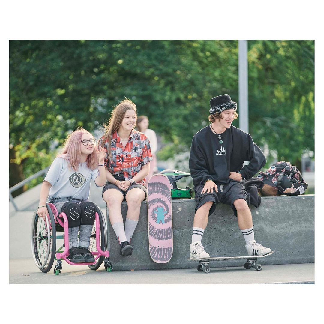 Three young people at a skatepark. Two girls sit together, one in a wheelchair, while a young man sits on a skateboard next to them.
