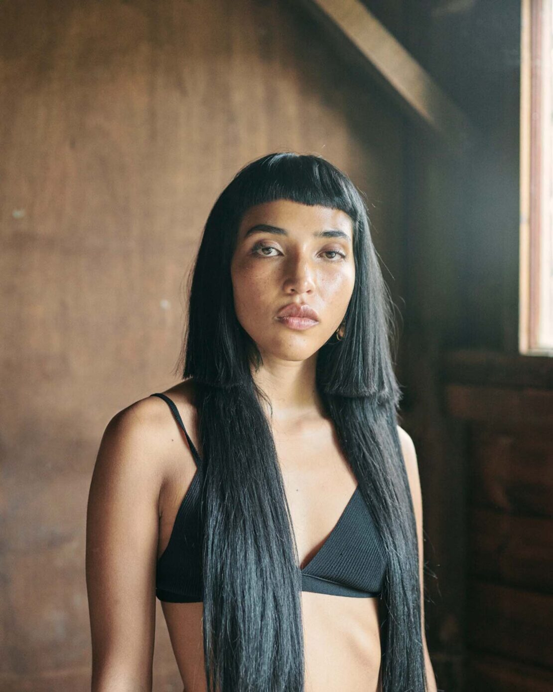 Portrait of a woman with long black hair in a black bikini top, posing indoors against a rustic wall with natural lighting.