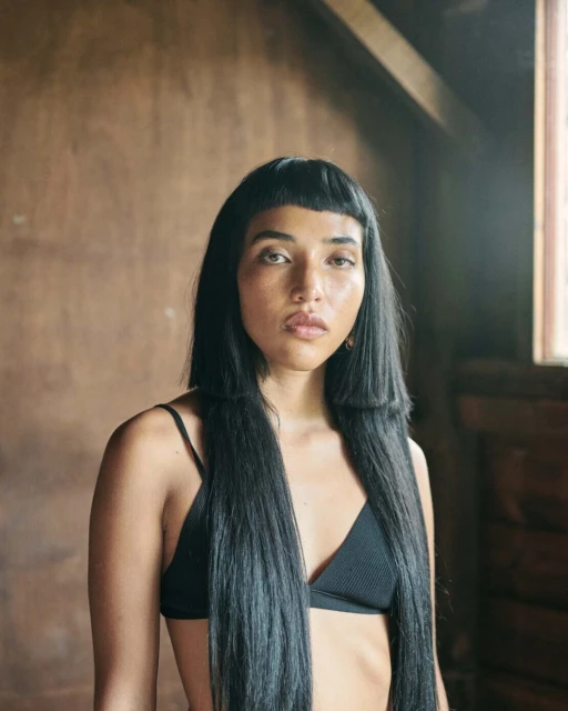 Portrait of a woman with long black hair in a black bikini top, posing indoors against a rustic wall with natural lighting.