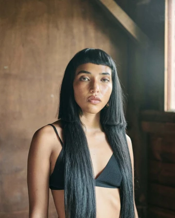 Portrait of a woman with long black hair in a black bikini top, posing indoors against a rustic wall with natural lighting.