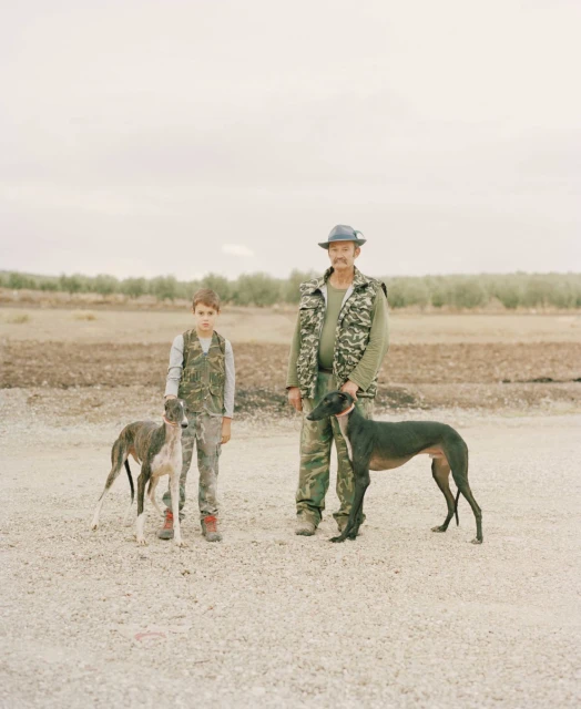 An adult man and a child, both wearing camouflage, stand with two greyhounds in an outdoor setting with sparse vegetation and an overcast sky.