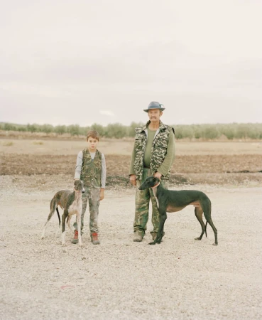 An adult man and a child, both wearing camouflage, stand with two greyhounds in an outdoor setting with sparse vegetation and an overcast sky.