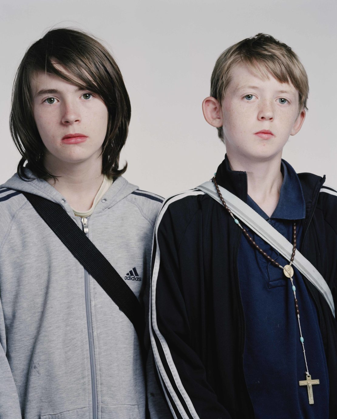 Two young boys stand indoors in a medium studio shot, one wearing a grey Adidas hooded sweatshirt and the other a blue track jacket.