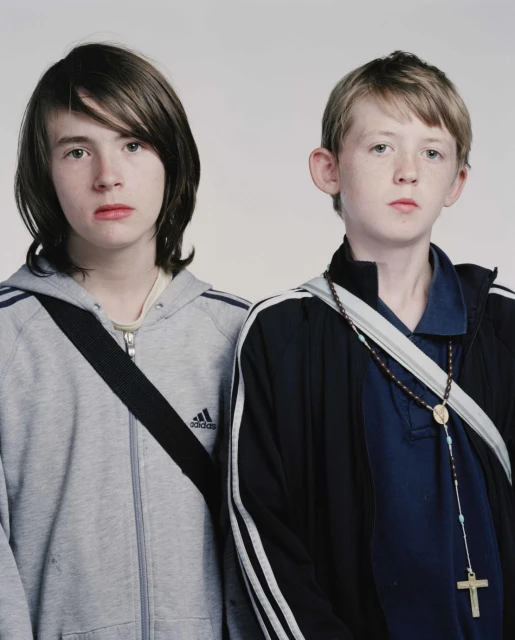 Two young boys stand indoors in a medium studio shot, one wearing a grey Adidas hooded sweatshirt and the other a blue track jacket.