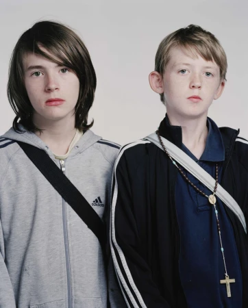 Two young boys stand indoors in a medium studio shot, one wearing a grey Adidas hooded sweatshirt and the other a blue track jacket.
