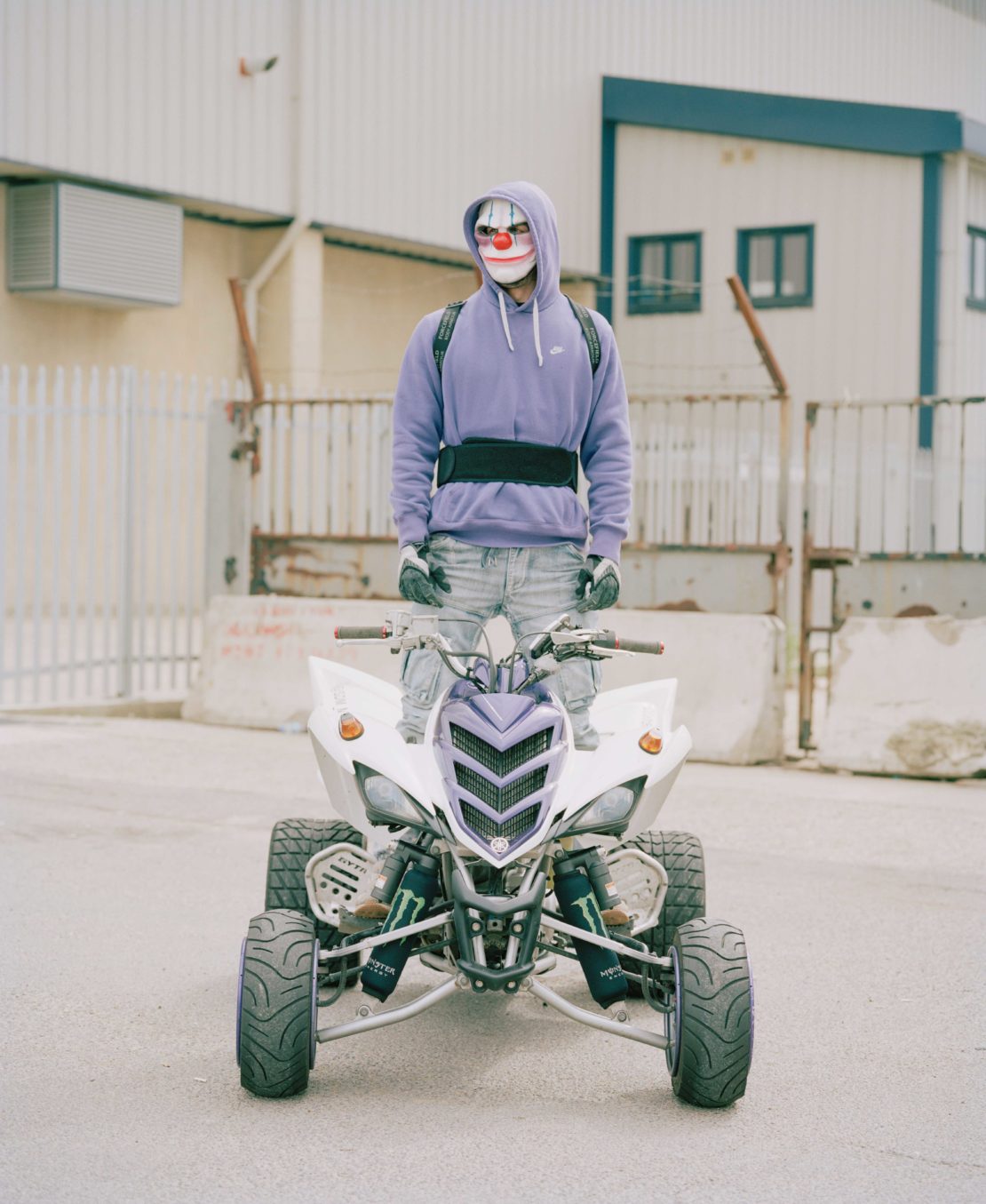 Person in a clown mask stands on a white ATV in an outdoor urban setting with muted colors and neutral light, wearing a purple hoodie.