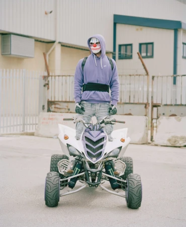 Person in a clown mask stands on a white ATV in an outdoor urban setting with muted colors and neutral light, wearing a purple hoodie.