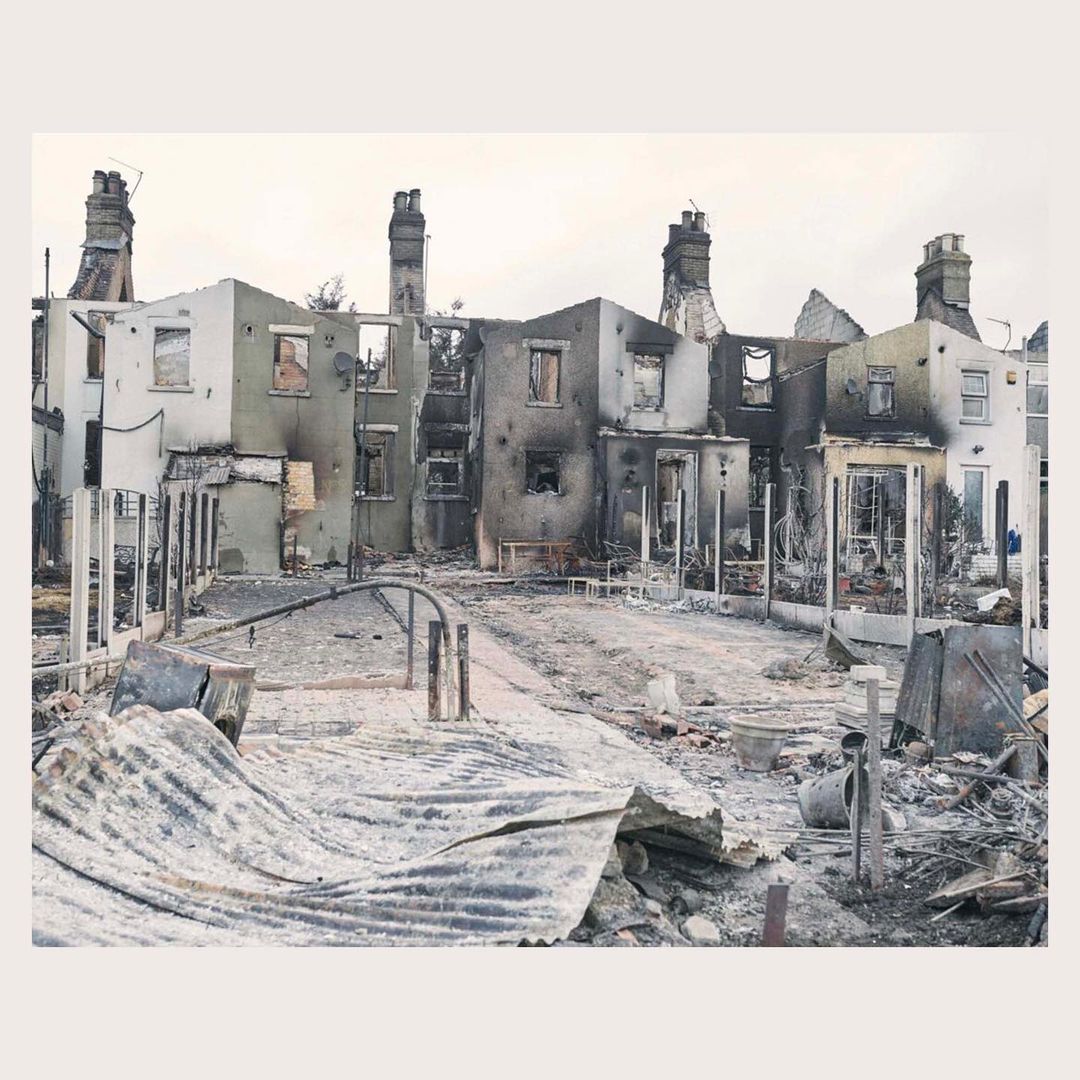 Destroyed row of houses with visible damage and debris in the aftermath of a fire, with chimneys still standing.