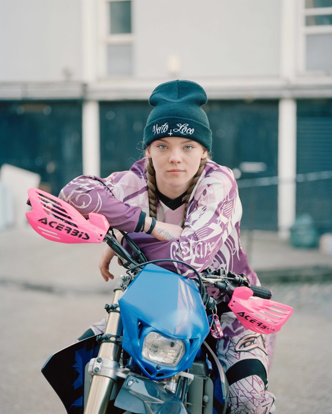 A young woman in motocross gear poses with her dirt bike on an overcast day, with urban buildings visible in the background.