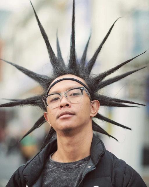 Person with spiked black hair wearing glasses and a black jacket in an outdoor urban setting with a blurred background, looking at camera.
