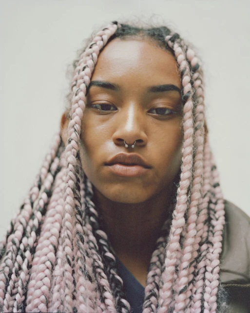 Portrait of a young adult woman with pink box braids and a septum piercing, facing forward in a close-up shot against a neutral background.