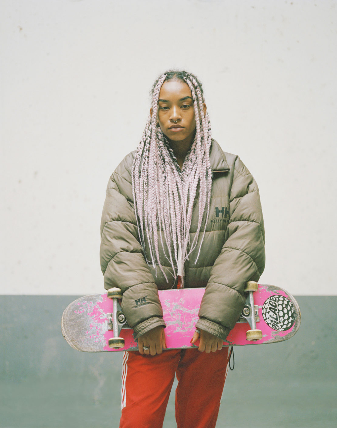 Young adult woman with pink-braided hair holds a pink skateboard while posing indoors against a concrete wall background. 