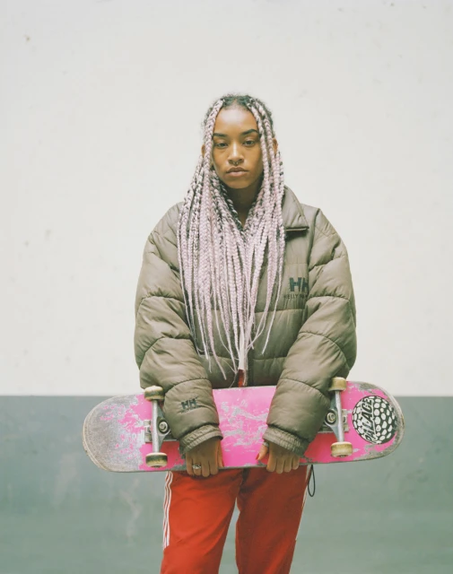 Young adult woman with pink-braided hair holds a pink skateboard while posing indoors against a concrete wall background. 