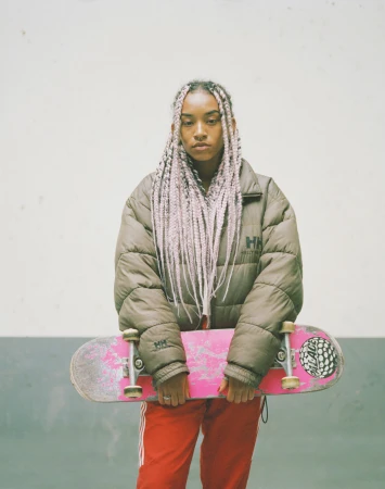 Young adult woman with pink-braided hair holds a pink skateboard while posing indoors against a concrete wall background. 
