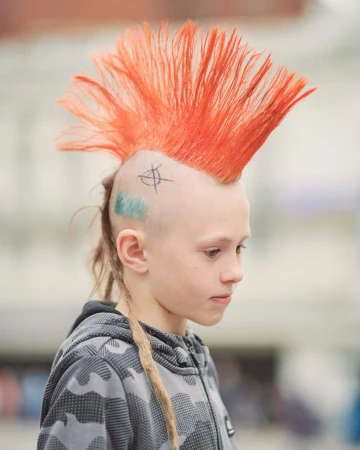 Young person with tall bright orange mohawk, shaved head tattoos, and a grey camouflage hoodie by Spencer Murphy