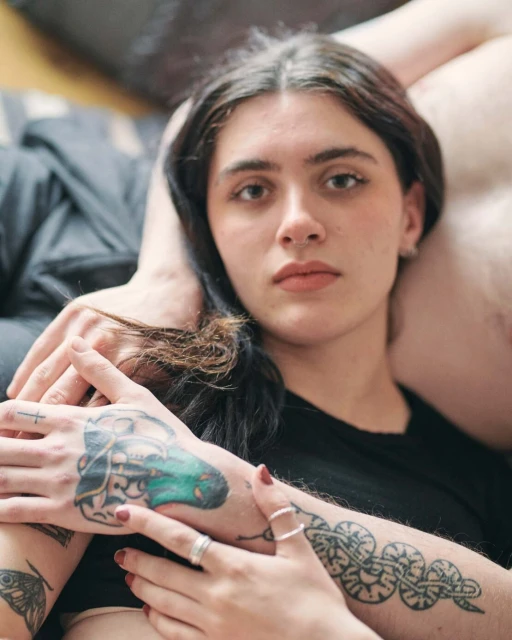 Close-up of a young woman with long brown hair, wearing a black t-shirt, lying down surrounded by arms with visible tattoos.