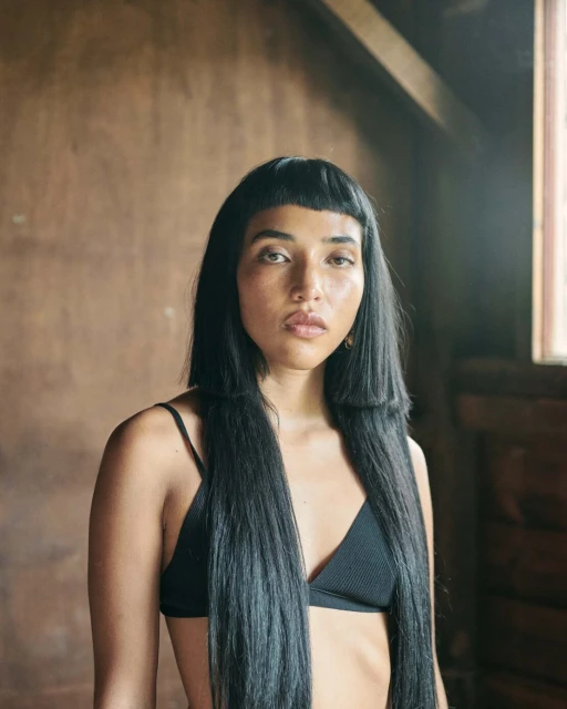 Close-up studio shot of a young woman with long black hair and a black top against a warm-toned background with window light.