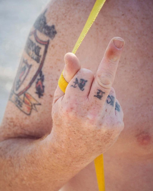 Torso with tattoos including Liverpool crest and hand with knuckle tattoos forming 'devil horns' gesture. Yellow strap is visible.