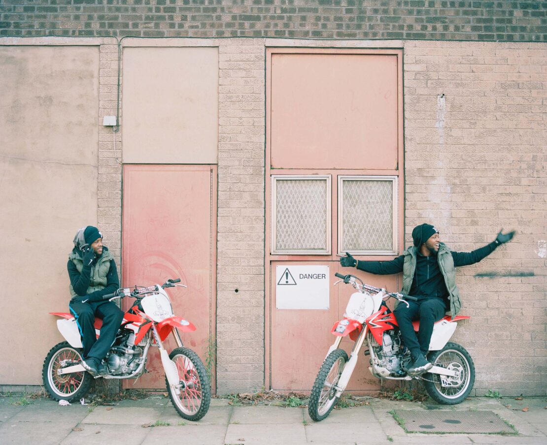 Two adult males sitting on red dirt bikes in front of a brick building with pale pink doors, wearing dark clothing and vests outdoors.