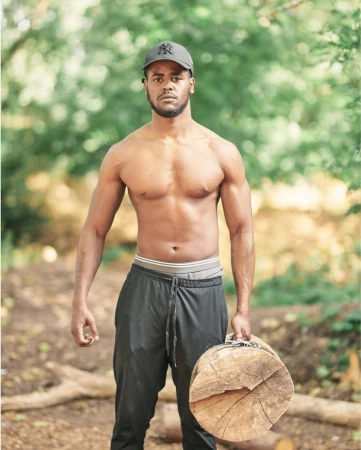 Shirtless man carrying a log in an outdoor setting, wearing a baseball cap and sweatpants, showcasing a fitness lifestyle in a forest.