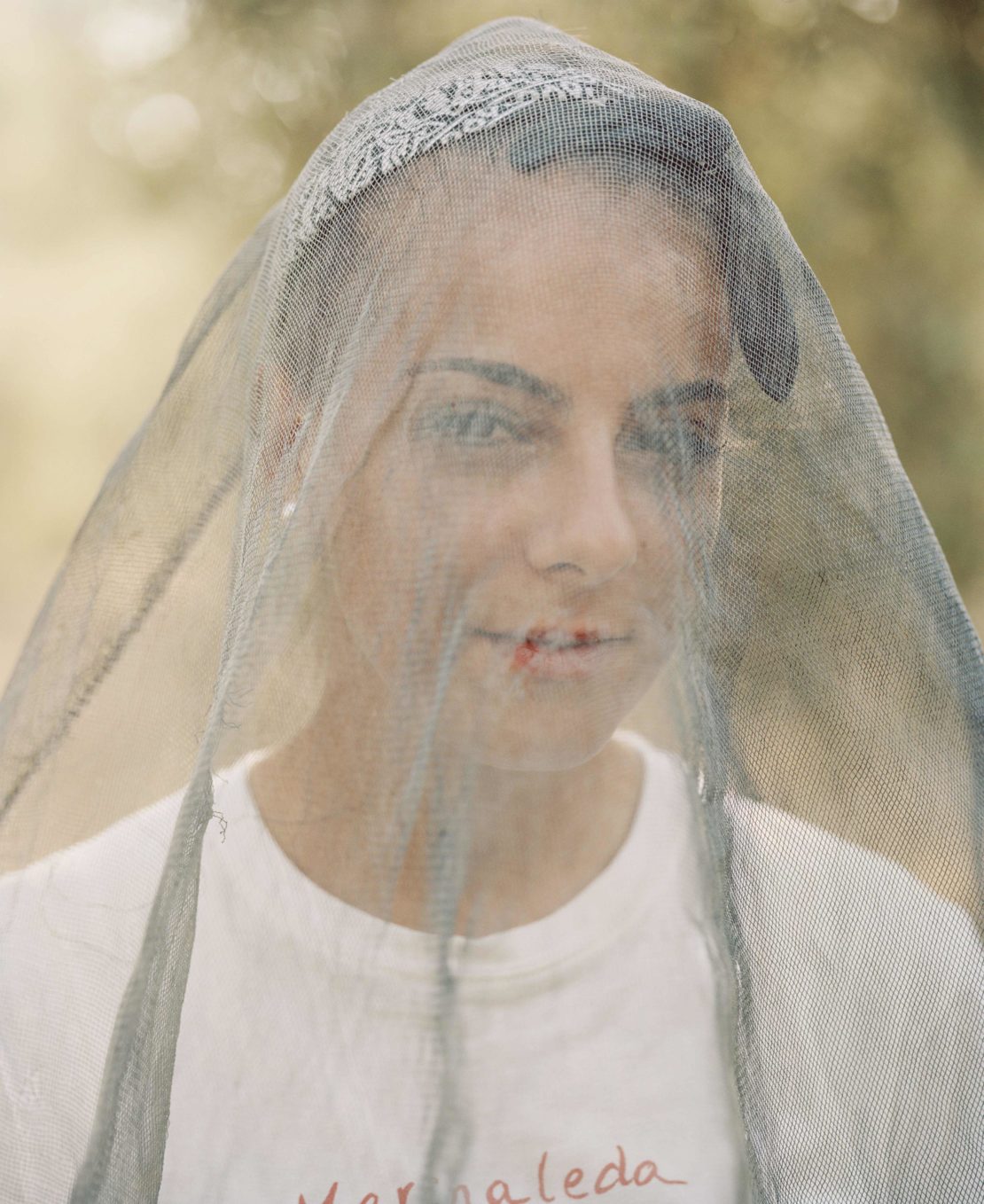 Portrait of a woman with a patterned bandana and a mesh veil standing outside in natural light. Close-up captures facial details.