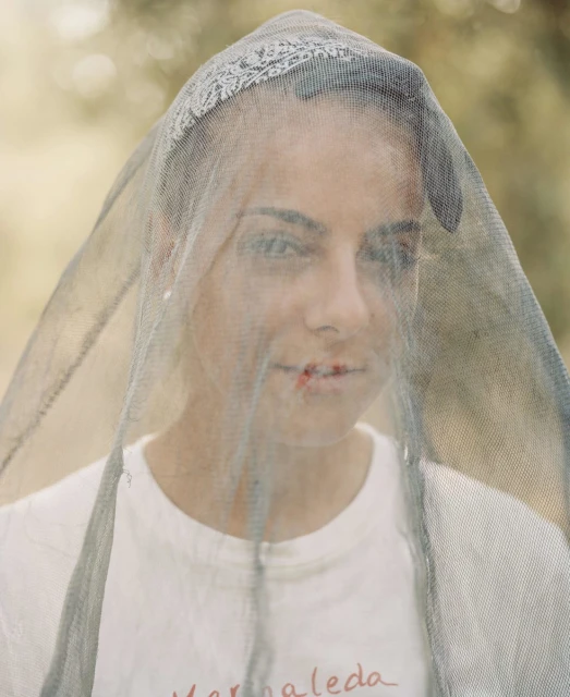 Portrait of a woman with a patterned bandana and a mesh veil standing outside in natural light. Close-up captures facial details.