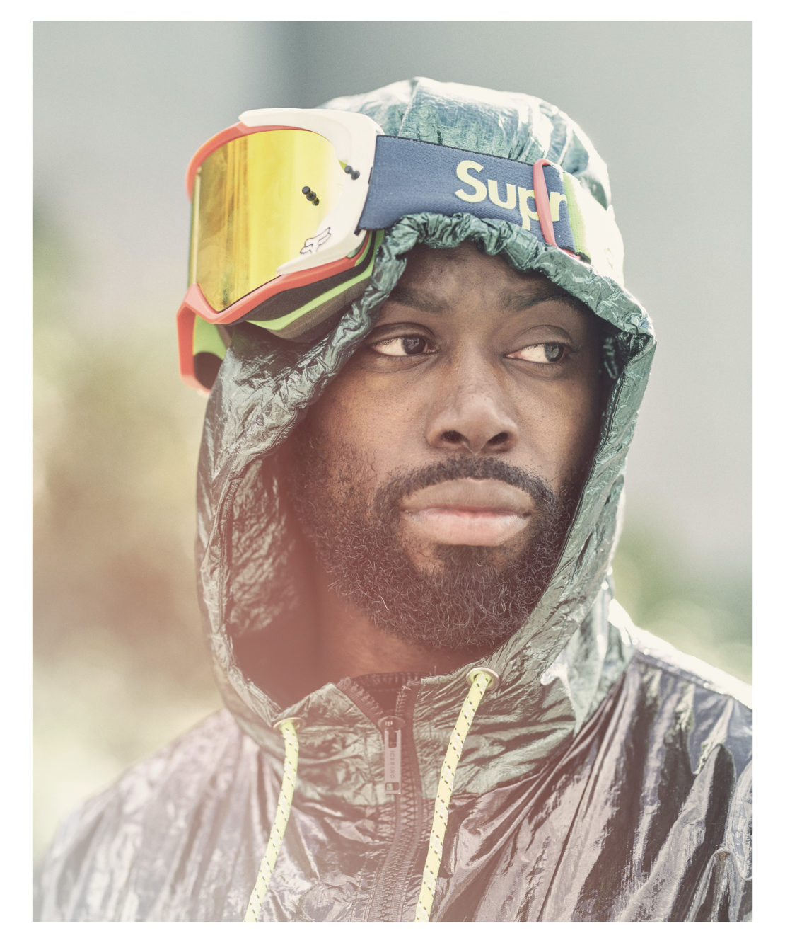 Portrait of a bearded Black adult male wearing ski goggles and a hooded, iridescent jacket with yellow drawstring.