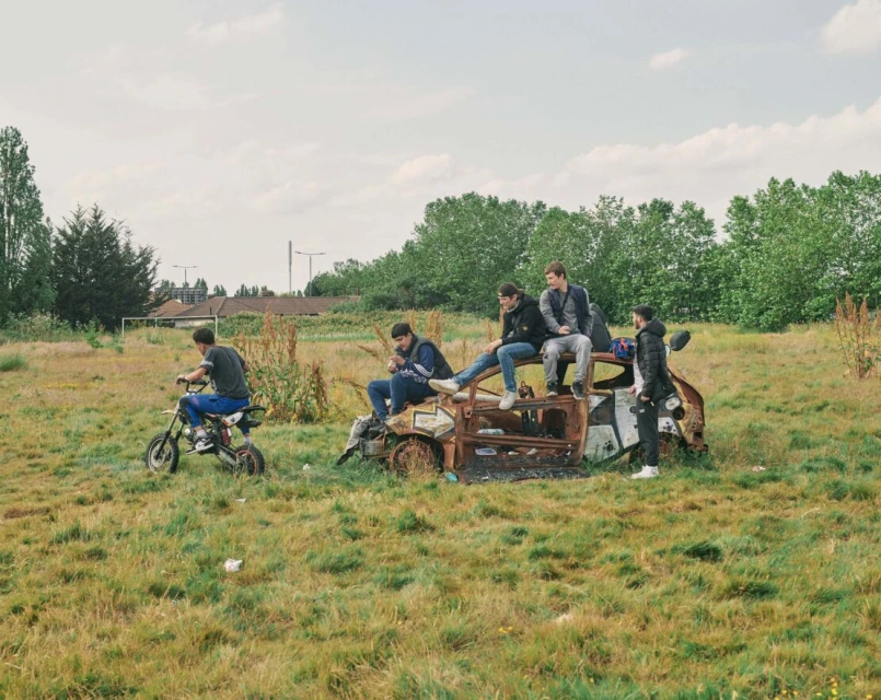 Group of young men on a dirt bike and sitting on a rusted car in a grassy field under a cloudy sky on a sunny day in a rural area.