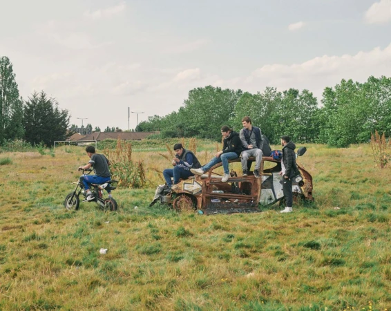 Group of young men on a dirt bike and sitting on a rusted car in a grassy field under a cloudy sky on a sunny day in a rural area.