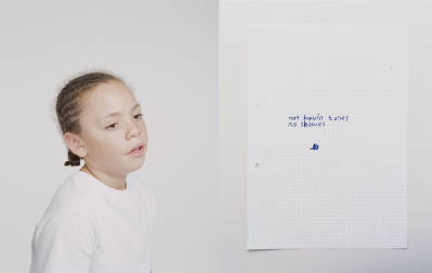 Young girl with braided hair looks at a sheet of paper with handwritten text against a neutral grey and white background.