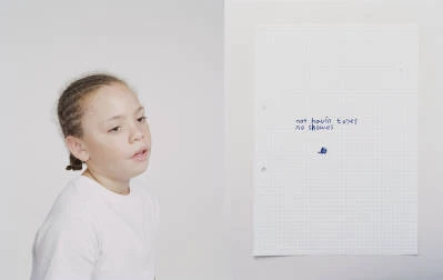 Young girl with braided hair looks at a sheet of paper with handwritten text against a neutral grey and white background.