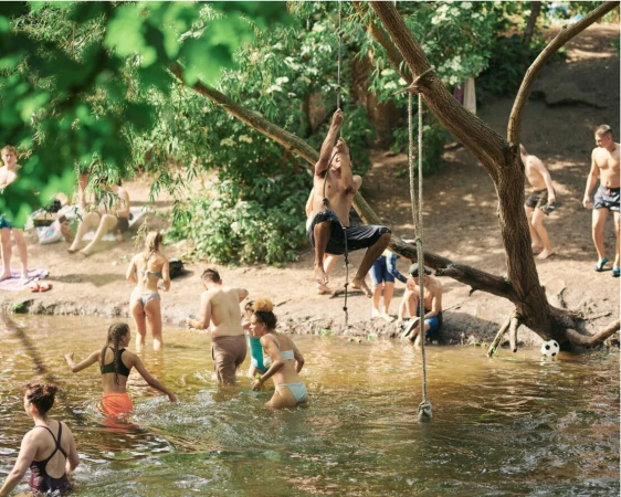Group of people swimming and playing in a lake surrounded by trees on a sunny day; one man climbs a rope swing.