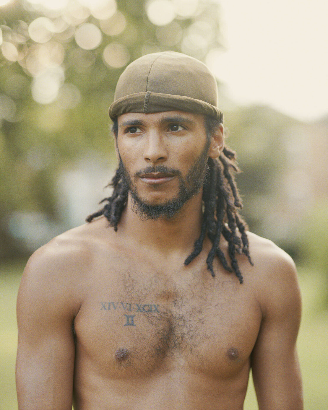 Close-up portrait of a shirtless adult male with dreadlocks and a do-rag outdoors in a natural setting with green foliage bokeh.