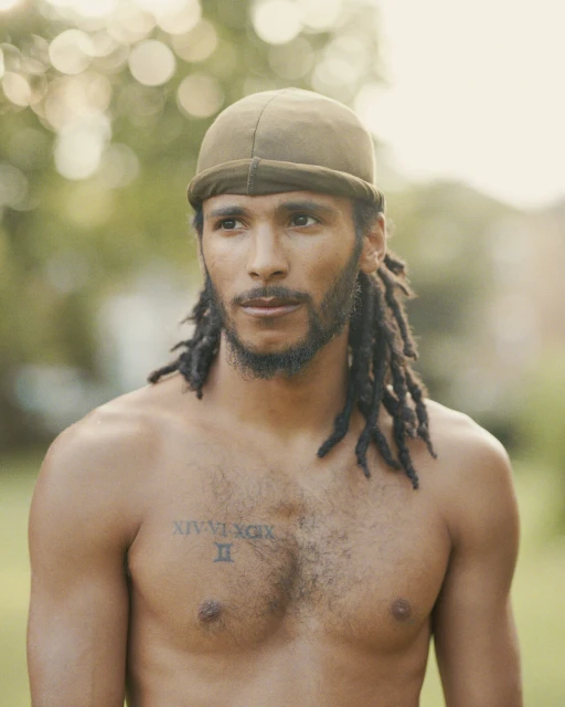 Close-up portrait of a shirtless adult male with dreadlocks and a do-rag outdoors in a natural setting with green foliage bokeh.