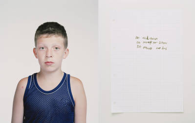 Portrait of a boy in a blue tank top, next to a handwritten note on lined paper against a white background in indoor lighting.
