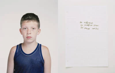 Portrait of a boy in a blue tank top, next to a handwritten note on lined paper against a white background in indoor lighting.