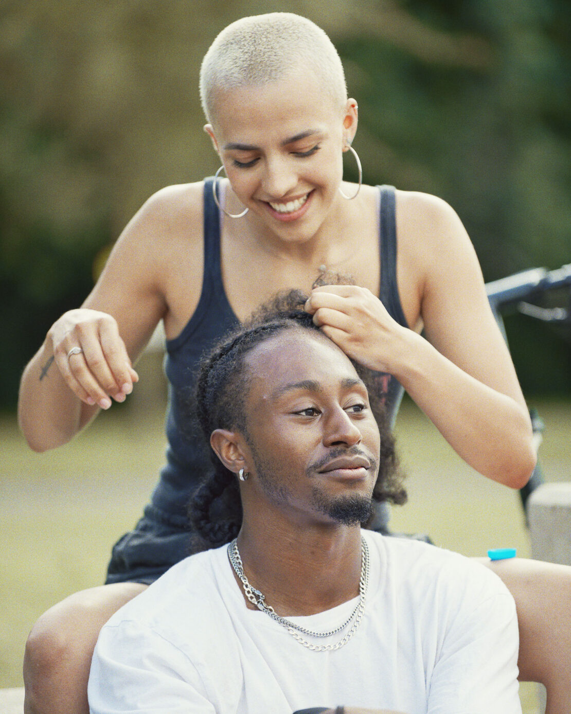 A smiling woman braids the hair of a man with locs outdoors on a sunny day, creating a warm and intimate moment in a natural setting.