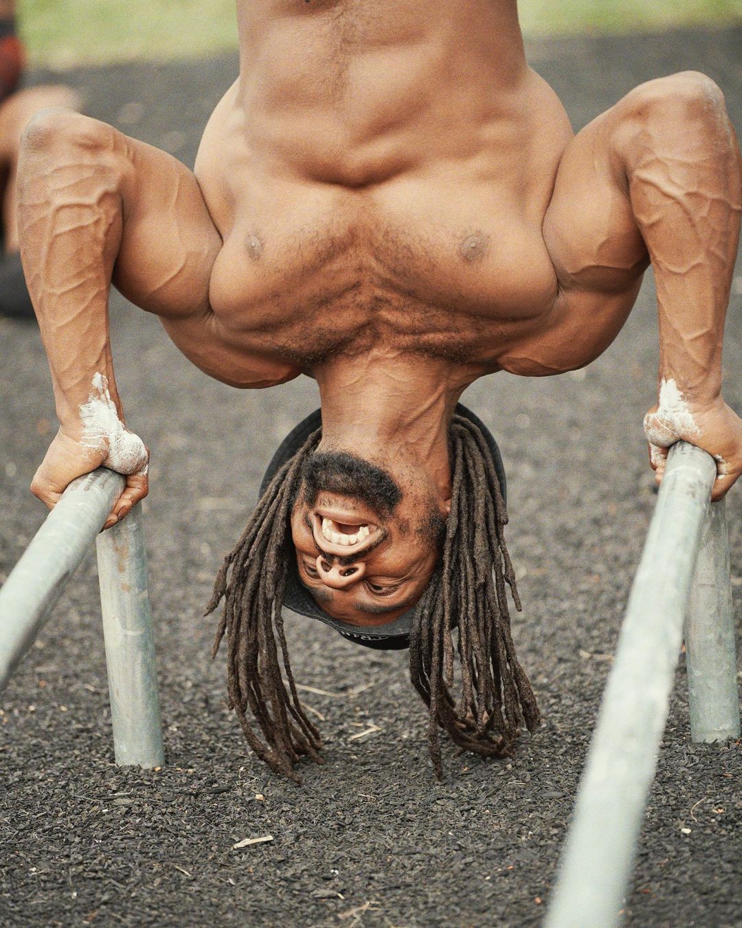 Muscular man with dreadlocks performs a handstand on parallel bars outdoors, showcasing strength and fitness in a dynamic pose.