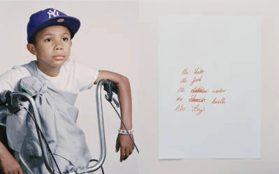 Young boy in baseball cap poses with bicycle next to red handwritten text on white paper, creating a contrast in style and message.