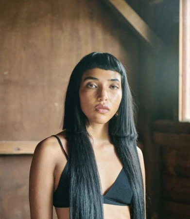 Portrait of a woman with long black hair and straight bangs wearing a black bralette inside a rustic wooden interior.
