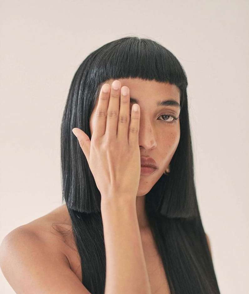 Striking portrait of a woman with a blunt bob hairstyle, covering one eye with her hand against a plain, light background in a studio setting.
