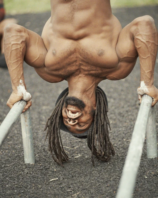 Muscular man performs an inverted handstand on parallel bars outdoors, showcasing impressive strength and physique.