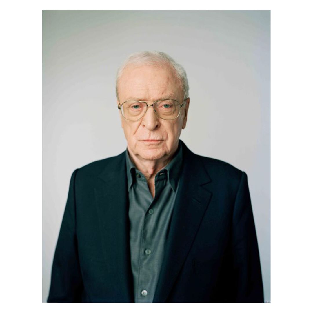 Close-up portrait of Michael Caine, an older gentleman with glasses wearing a gray shirt and a black suit jacket against a light backdrop.
