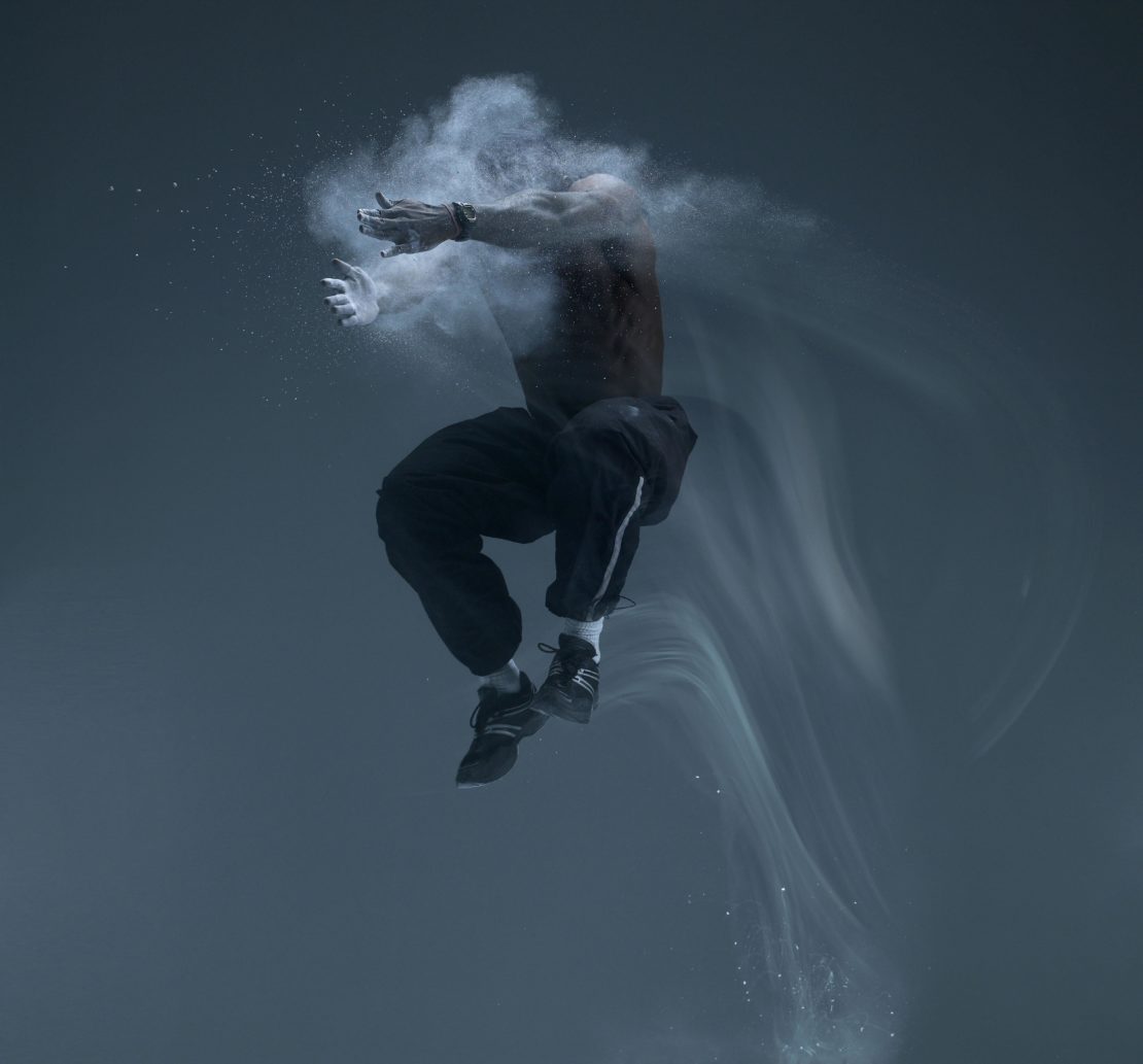 Athletic man mid-air, shirtless in black pants and shoes, surrounded by white powder against a gray background, studio lighting, action shot.